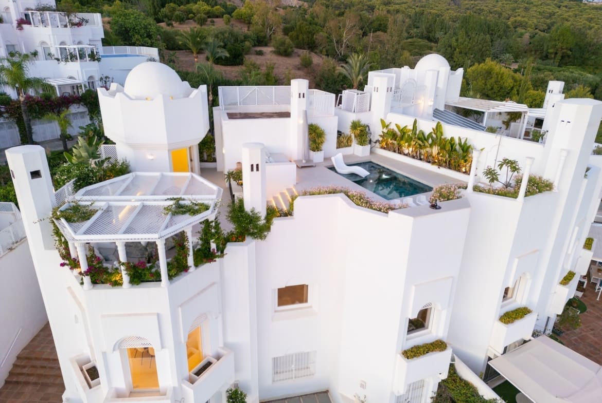 Aerial view of a white Mediterranean villa complex with a rooftop pool, terraces, and lush greenery on a hillside.