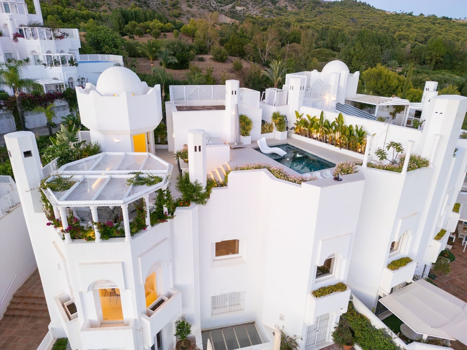 Aerial view of a white Mediterranean villa complex with a rooftop pool, terraces, and lush greenery on a hillside.