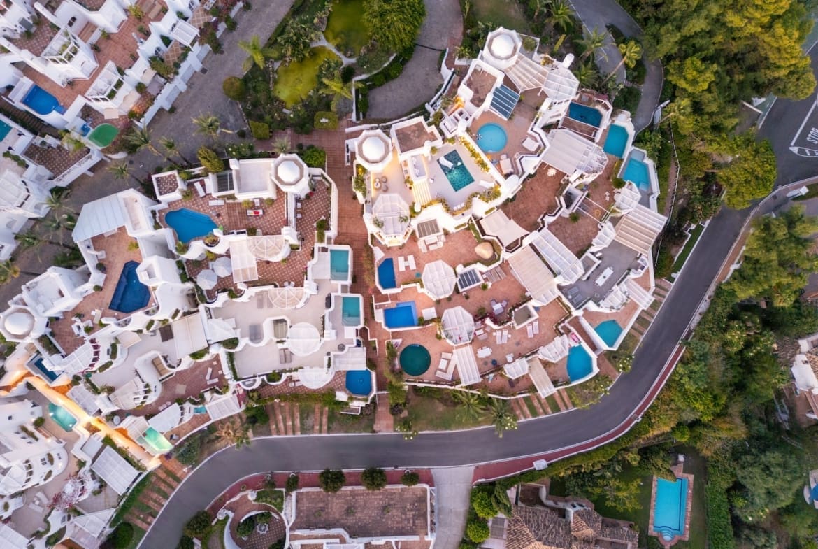 Aerial view of a white-walled resort with many blue pools, terracotta patios, and palm trees sprawled across a hillside.