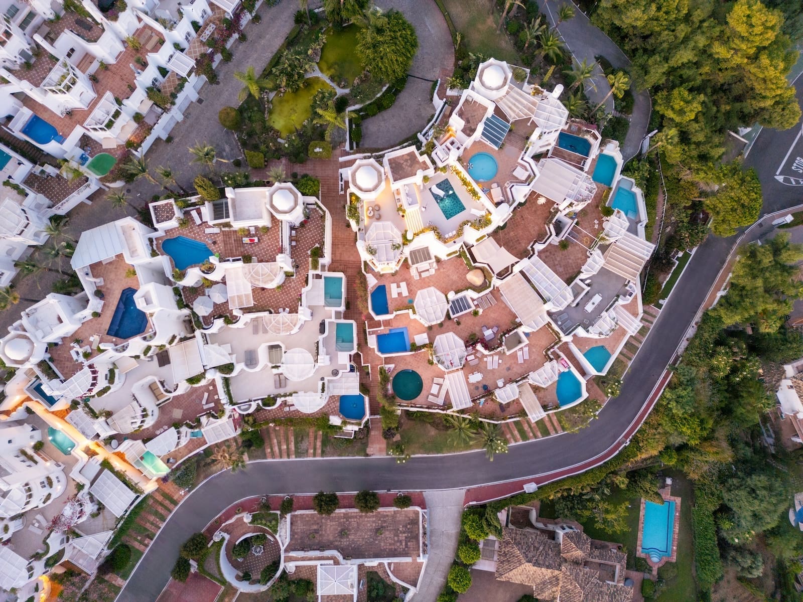 Aerial view of a white-walled resort with many blue pools, terracotta patios, and palm trees sprawled across a hillside.