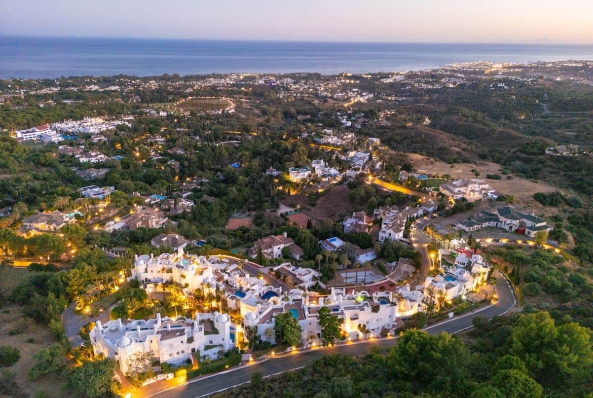 Aerial view of a coastal hillside residential neighborhood at dusk with illuminated streets and the ocean in the background.