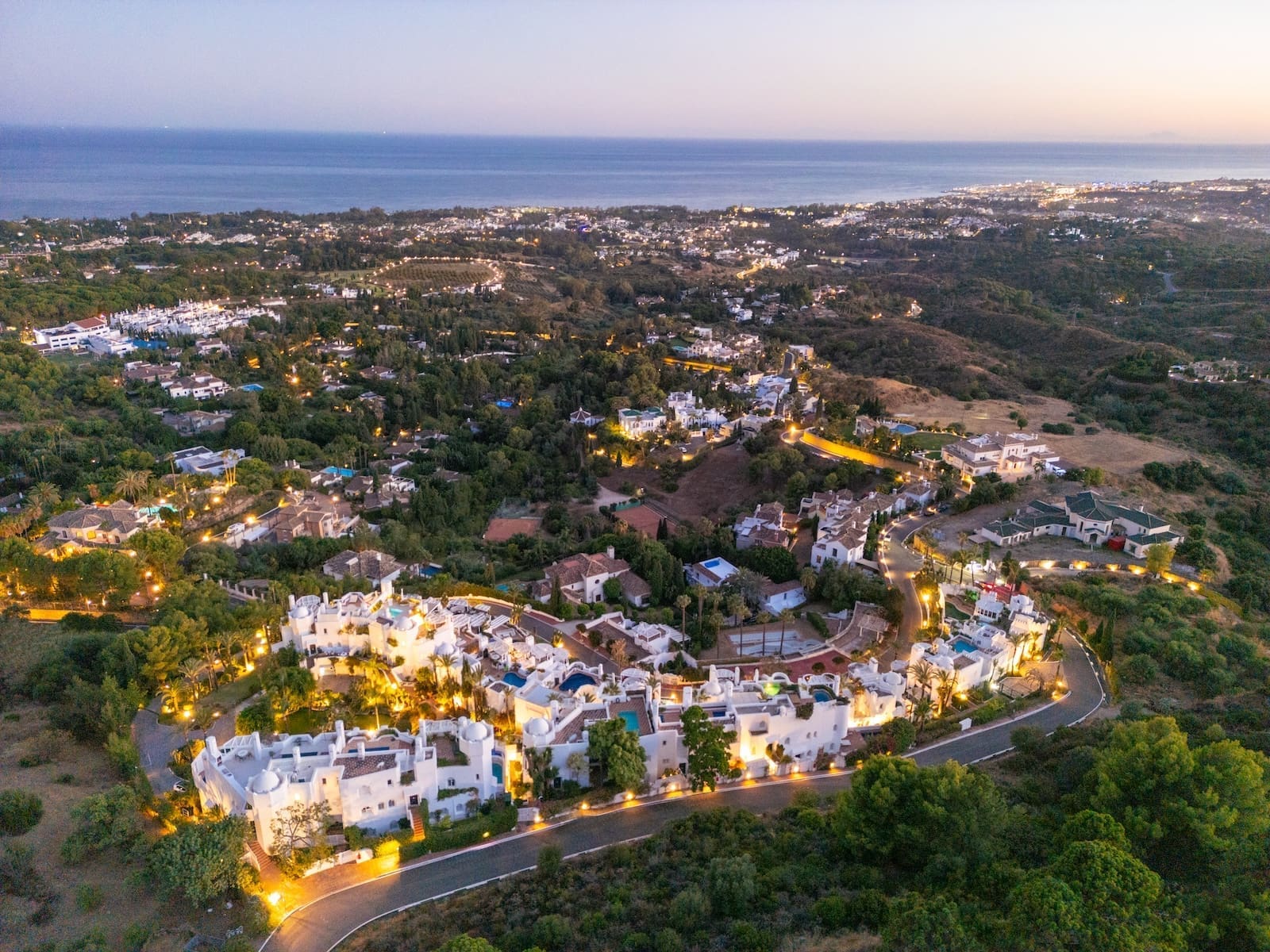 Aerial view of a coastal hillside residential neighborhood at dusk with illuminated streets and the ocean in the background.