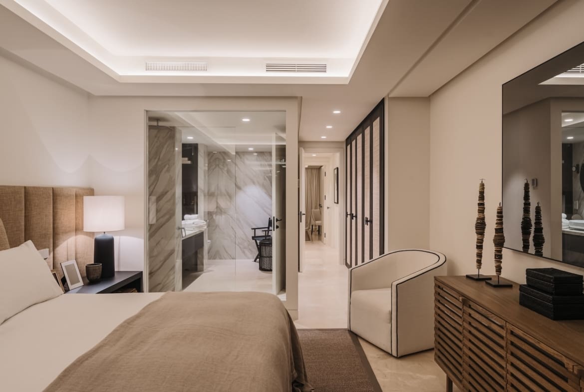 Hotel bedroom with king bed, beige headboard, and a glass-enclosed bathroom to the left.