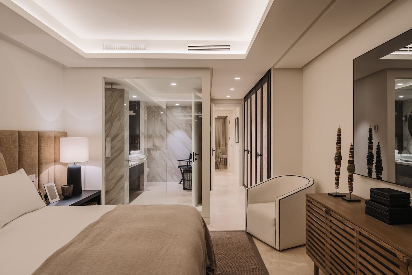 Hotel bedroom with king bed, beige headboard, and a glass-enclosed bathroom to the left.