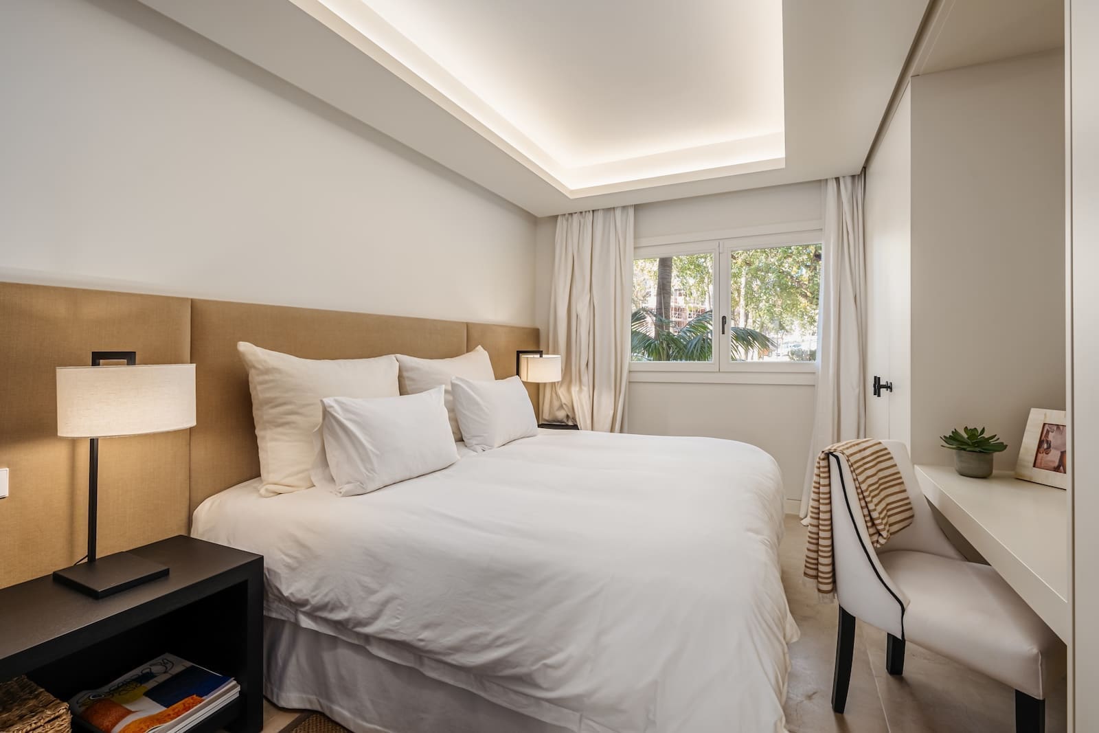 Bright hotel bedroom with a white-bedding bed, beige upholstered headboard, two nightstands and lamps, and a window with sheer curtains.