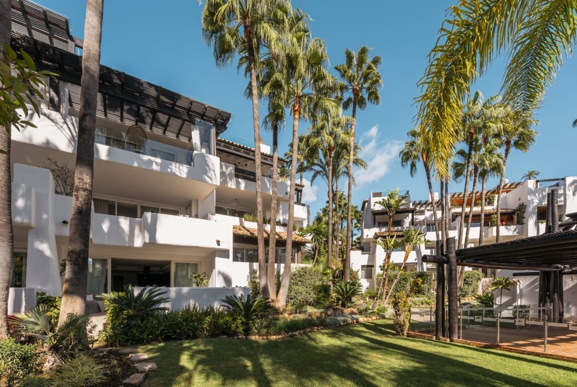 White modern resort buildings with balconies set among tall palm trees under a bright blue sky.