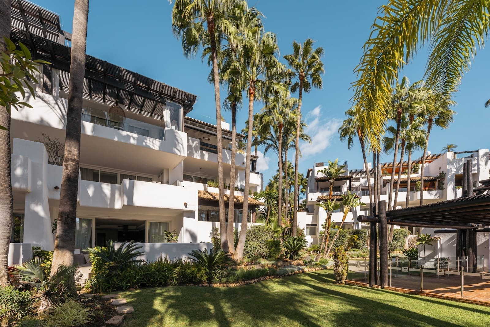White modern resort buildings with balconies set among tall palm trees under a bright blue sky.