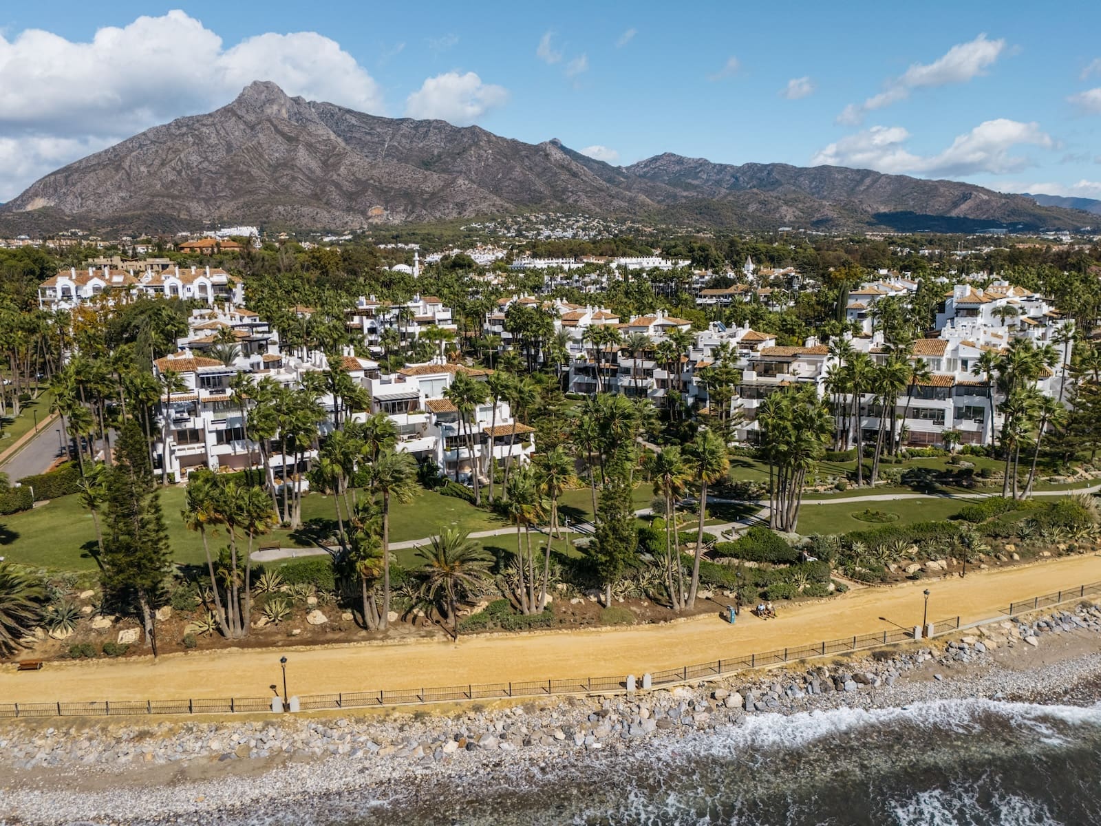Aerial view of a white seaside resort with palm trees, a sandy beach, and a rocky shoreline set against a mountain backdrop.