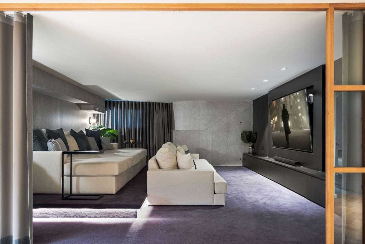 Modern basement living room with a beige sectional, dark throw pillows, and a large wall-mounted TV on a dark accent wall.