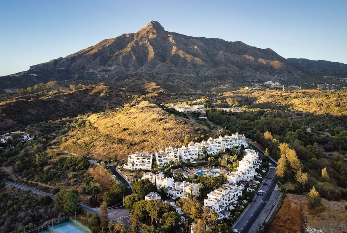 Aerial view of white apartment buildings clustered on a hillside with a mountain in the background and a pool center.