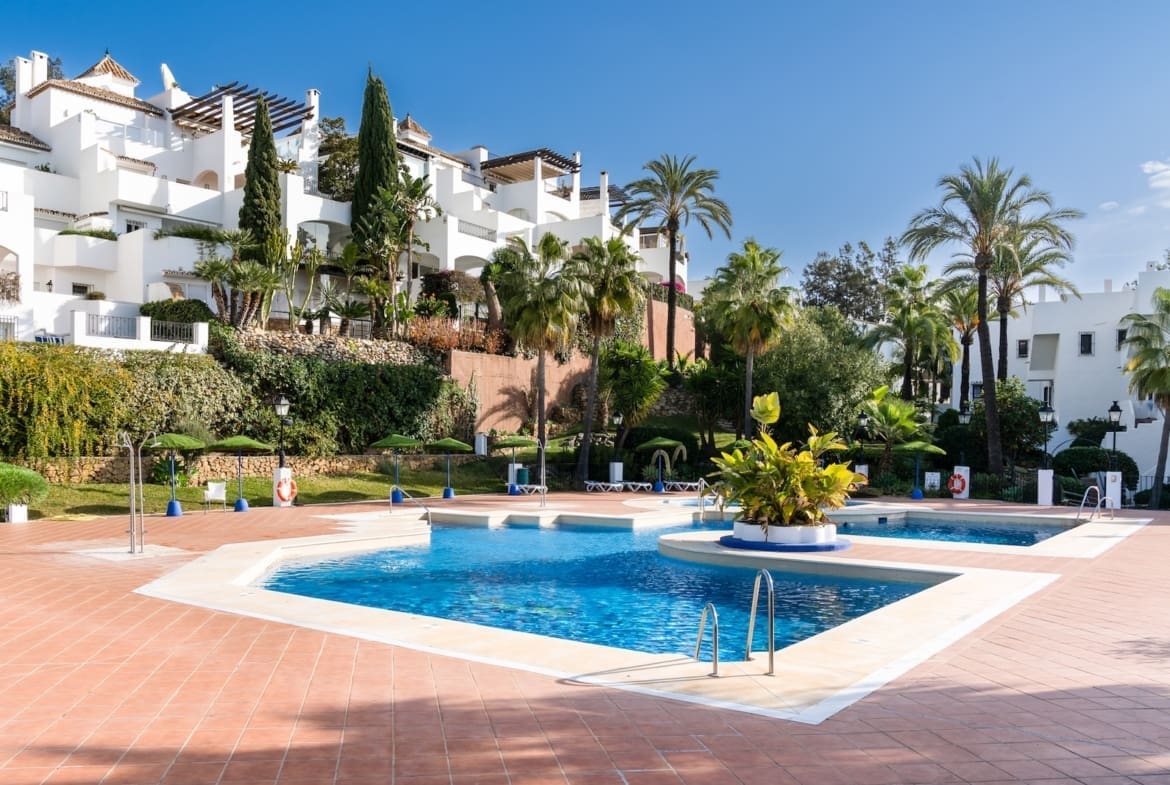 Resort pool area with blue water, terracotta tiles, and white Mediterranean-style buildings surrounded by palm trees on a sunny day.
