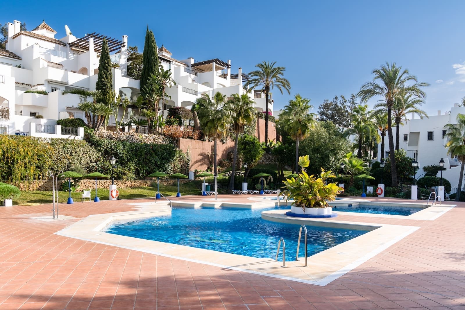 La Maison Saint Germain luxury townhouse Golden Mile Marbella – MDR Luxury Homes 45 Resort pool area with blue water, terracotta tiles, and white Mediterranean-style buildings surrounded by palm trees on a sunny day.