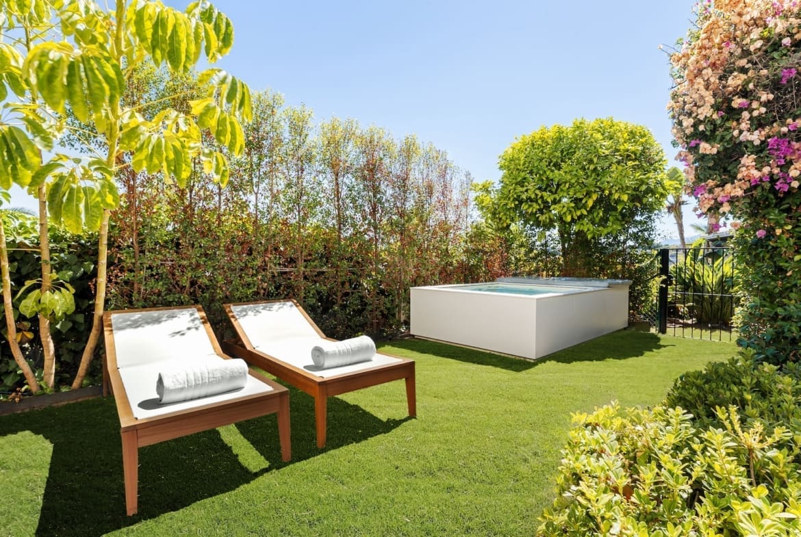 Two wooden sun loungers with white cushions and rolled towels on a green lawn in a sunny garden beside a raised rectangular pool.