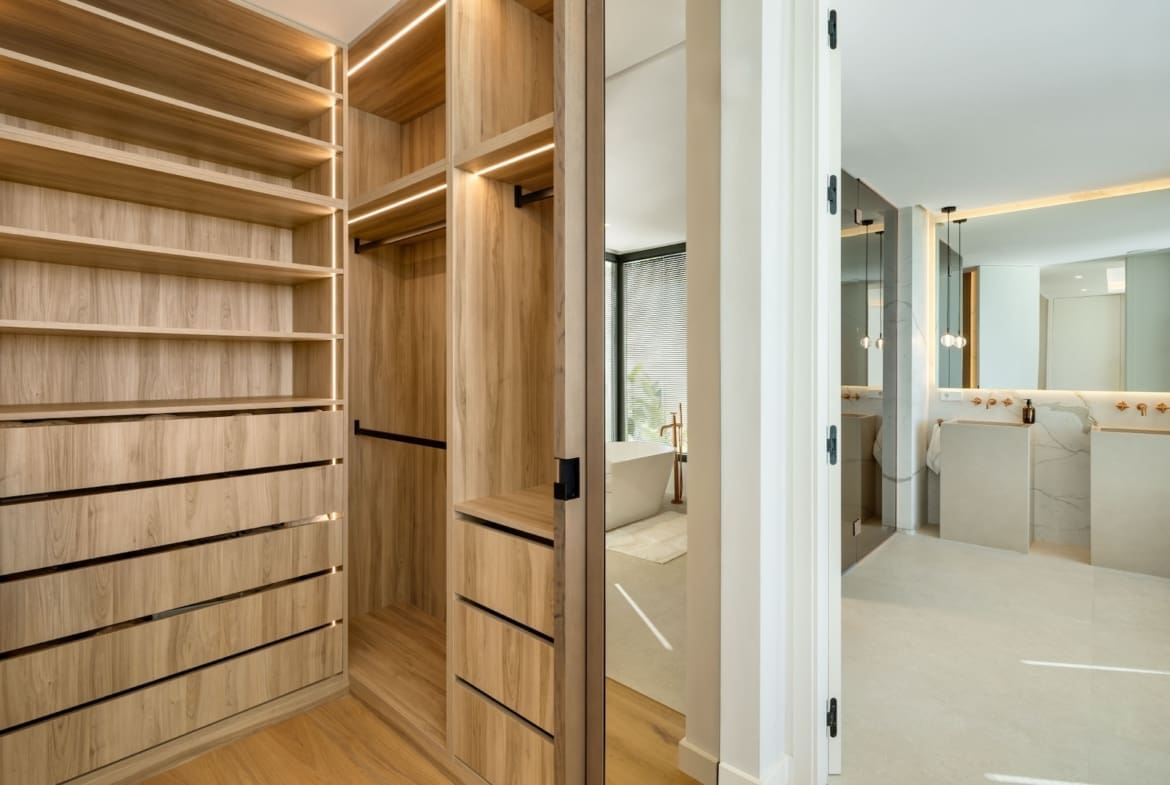 Walk-in closet with light wood shelves and drawers, opening to a bright marble bathroom with a freestanding tub.