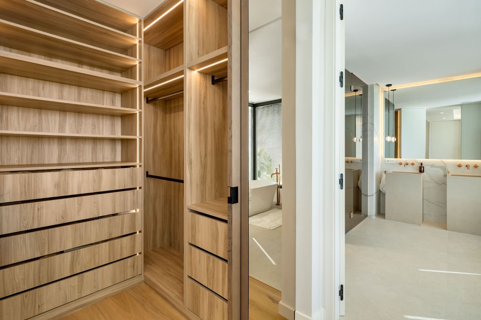 Walk-in closet with light wood shelves and drawers, opening to a bright marble bathroom with a freestanding tub.