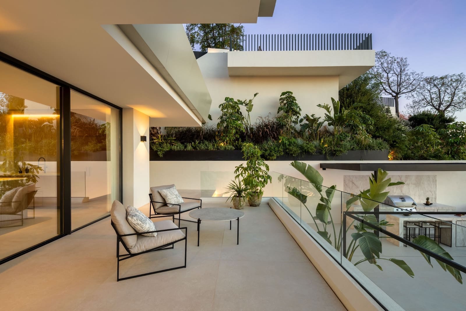 Modern outdoor terrace with two cushioned chairs and a round table, glass railing, and lush planters; interior kitchen visible through glass doors.