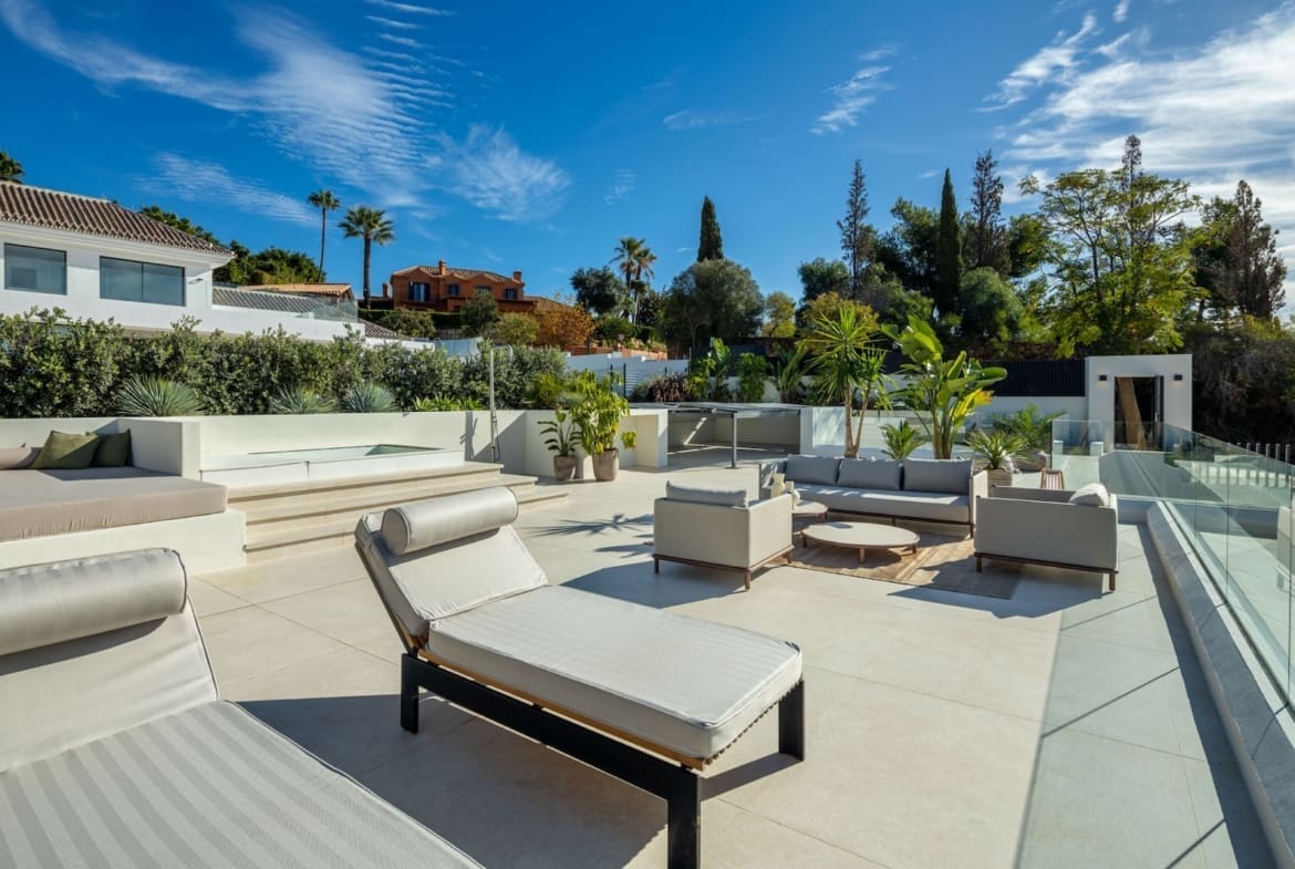 Modern outdoor lounge on a sunlit terrace with white cushioned seating, glass railing, and tropical plants under a blue sky.