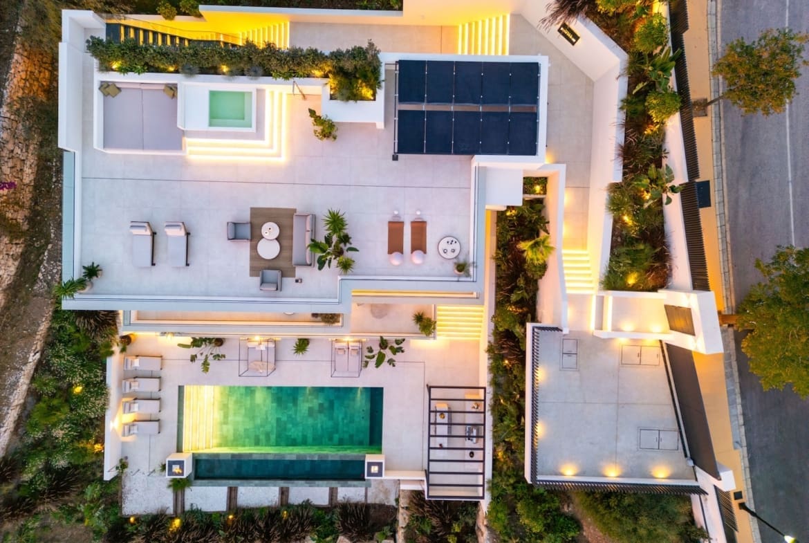 Aerial view of a modern white villa with a rectangular pool, outdoor lounge, sunbeds, and surrounding terraces at dusk.