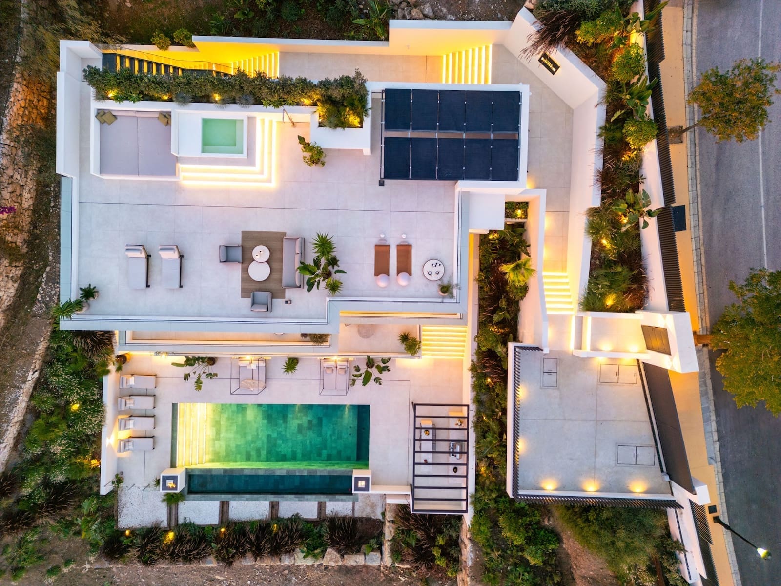Aerial view of a modern white villa with a rectangular pool, outdoor lounge, sunbeds, and surrounding terraces at dusk.