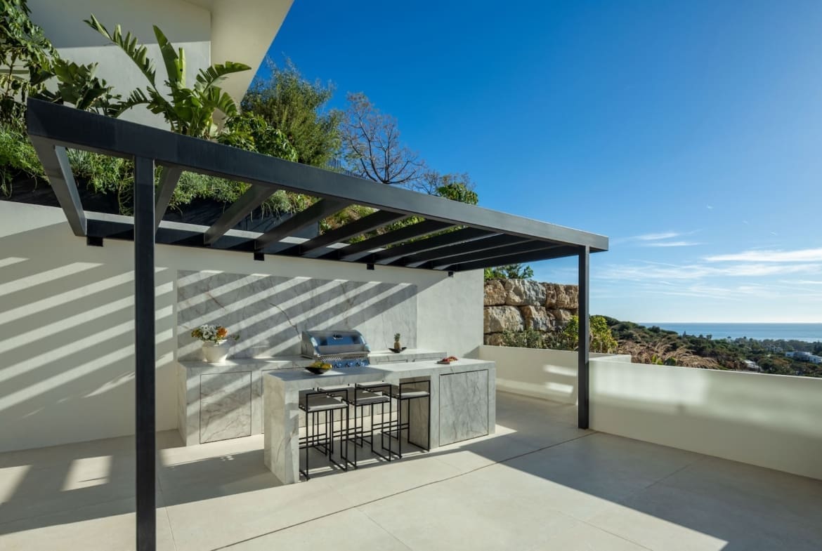 Outdoor kitchen with marble counters and a built-in grill under a black pergola, overlooking a coastal valley and ocean on a sunny day
