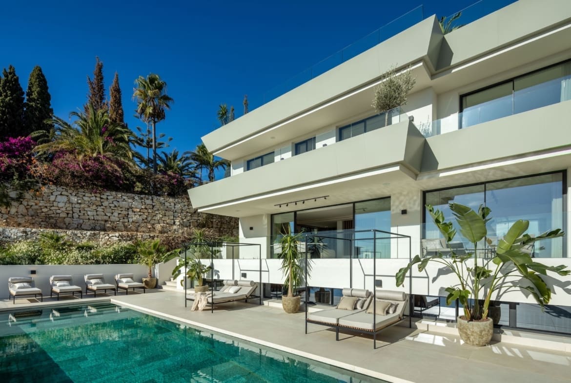 Modern villa with a rectangular pool, white patio furniture, and glass railings under a bright blue sky.