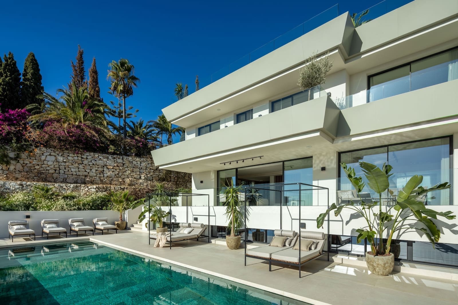 Modern villa with a rectangular pool, white patio furniture, and glass railings under a bright blue sky.