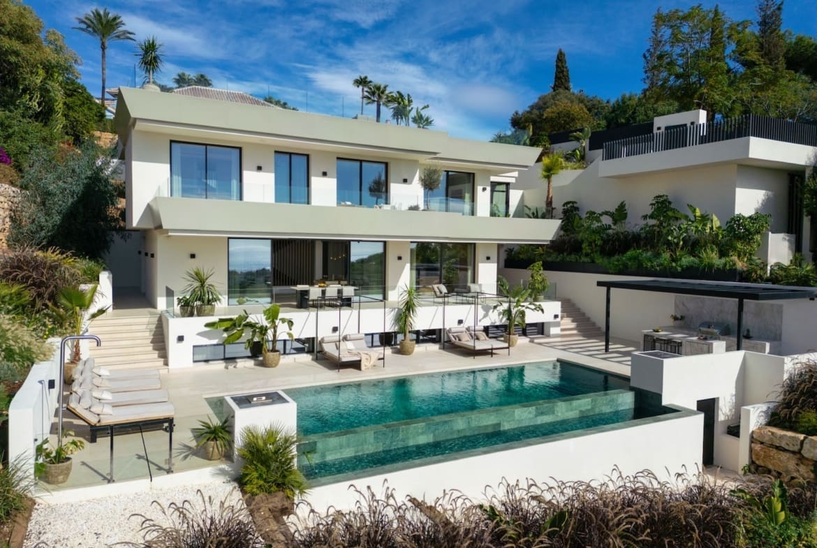 Modern white villa with expansive glass doors, multiple terraces, and a turquoise rectangular pool under a blue sky.