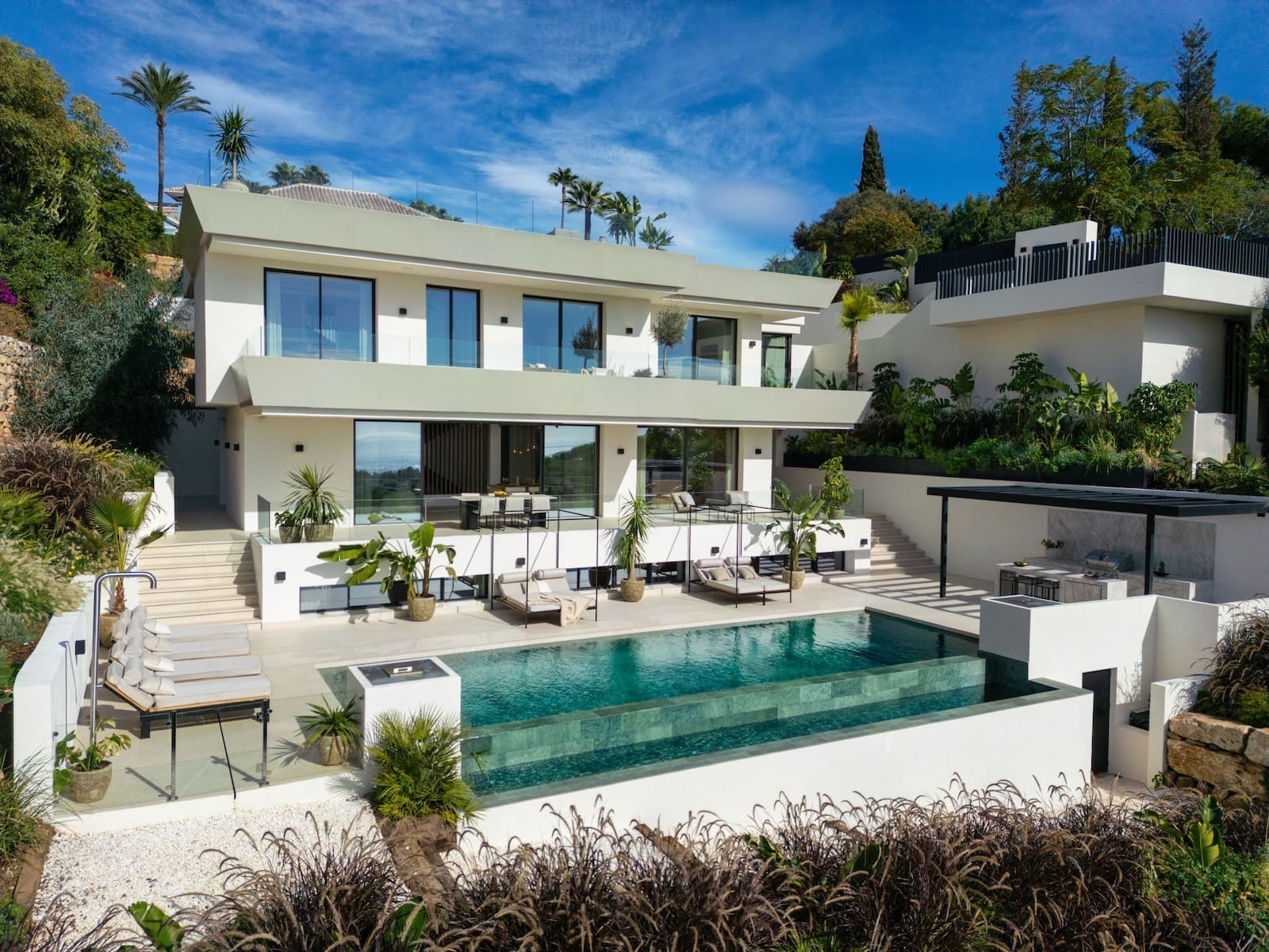 Modern white villa with expansive glass doors, multiple terraces, and a turquoise rectangular pool under a blue sky.