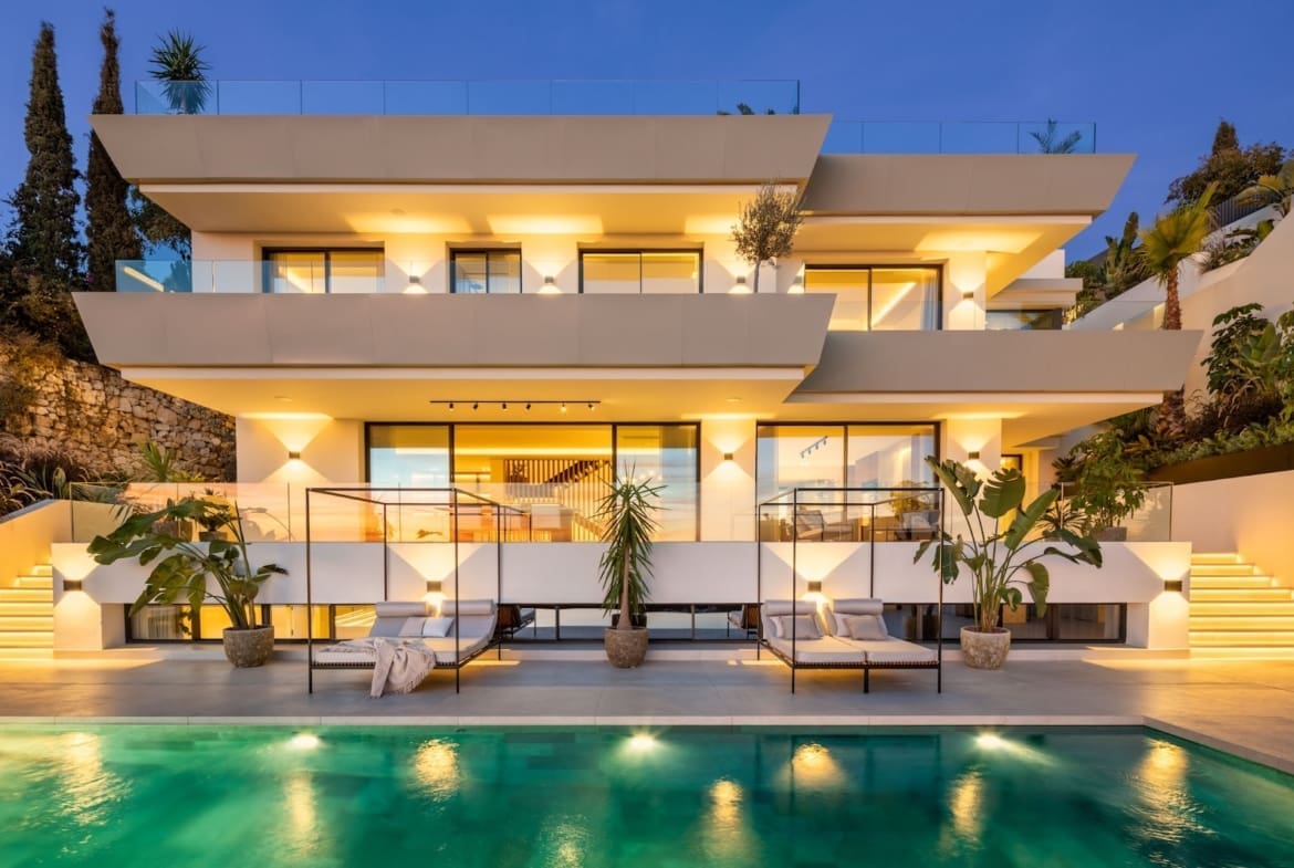 Modern multi-level white villa with glass balconies illuminated at dusk, adjacent to a turquoise swimming pool and lounge chairs.
