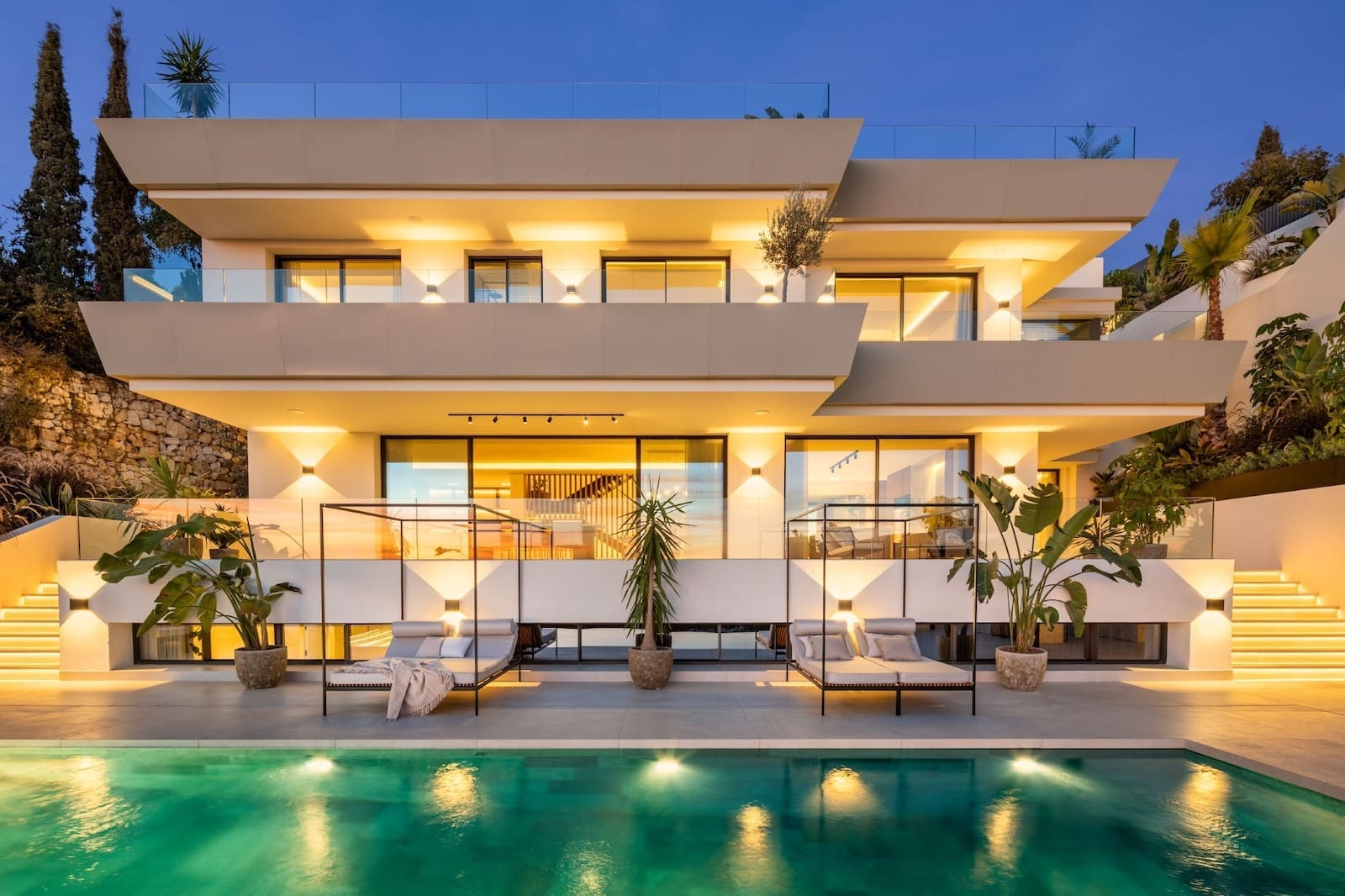 Modern multi-level white villa with glass balconies illuminated at dusk, adjacent to a turquoise swimming pool and lounge chairs.