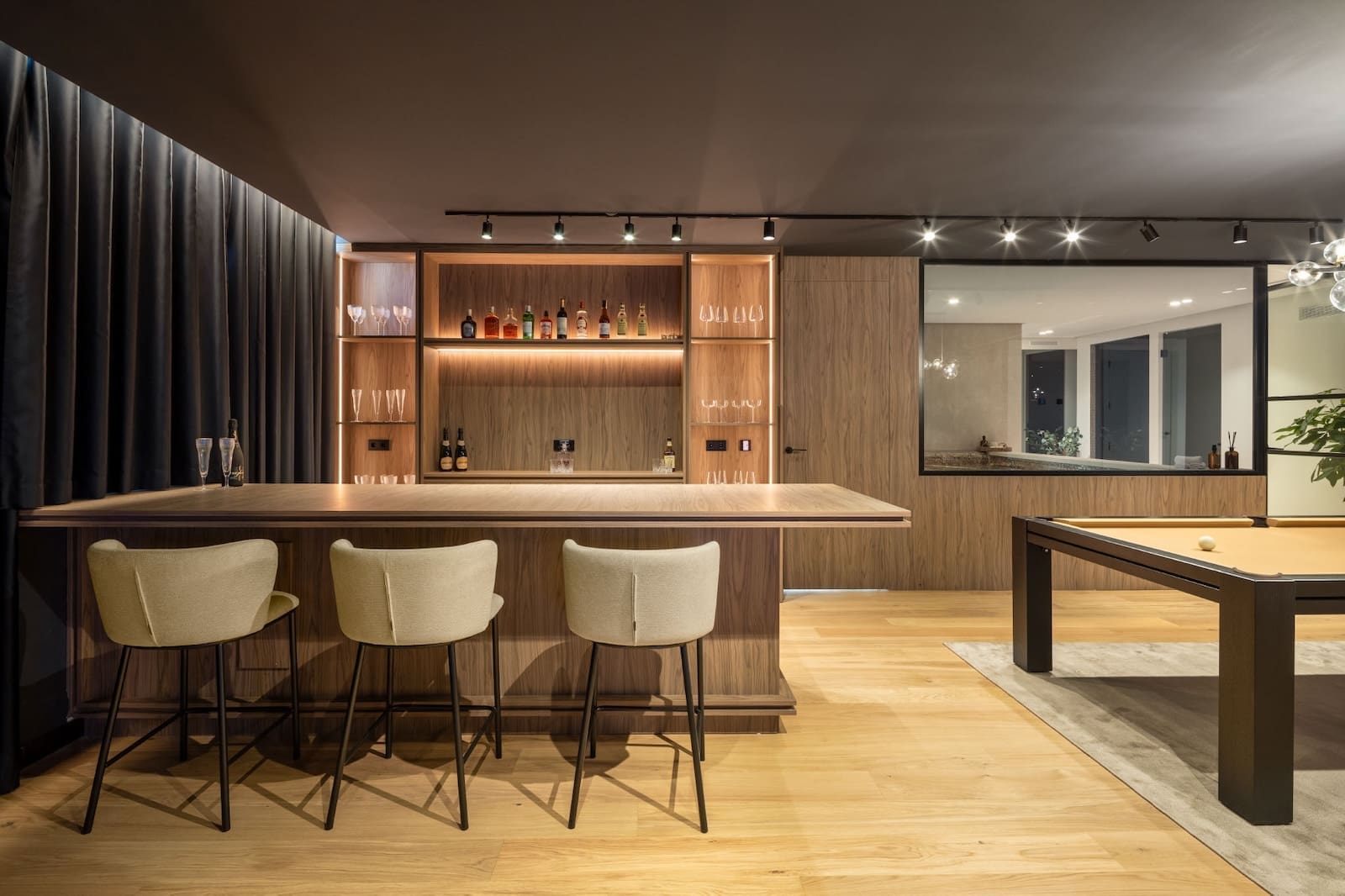 Modern wood bar with backlit shelves, bottles and glassware, three beige bar stools; pool table visible to the right.