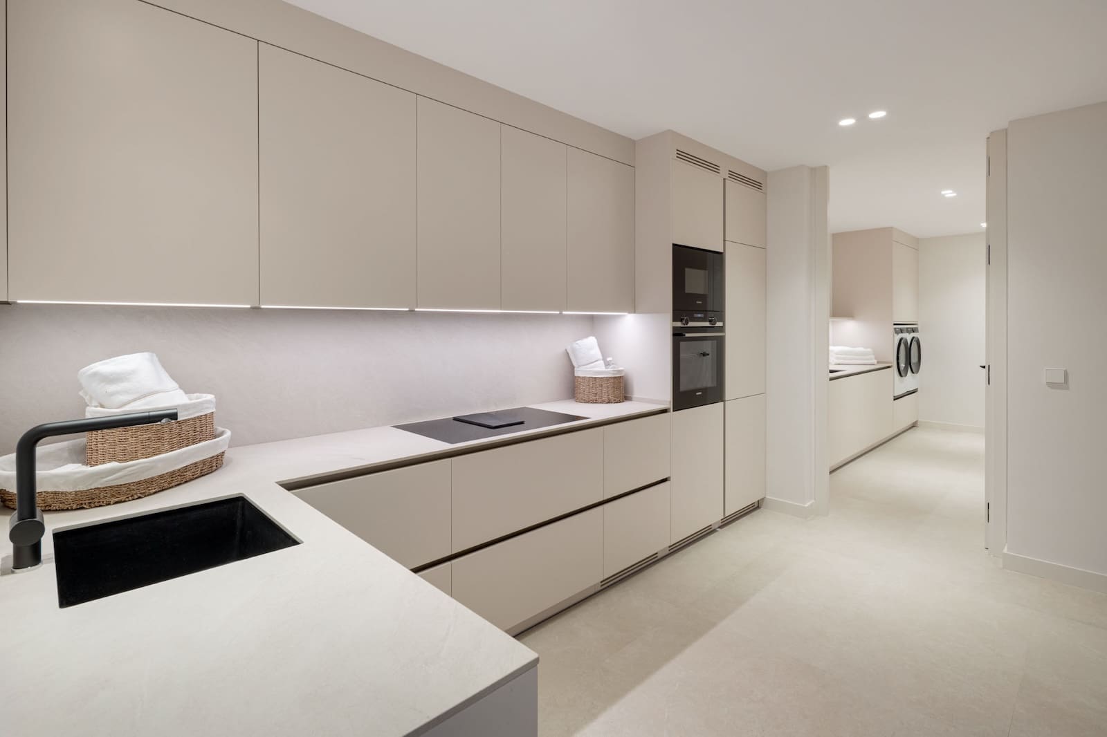 Minimalist beige kitchen with long countertops, handle-less cabinets, and built-in oven and microwave. Under-cabinet lighting adds glow.