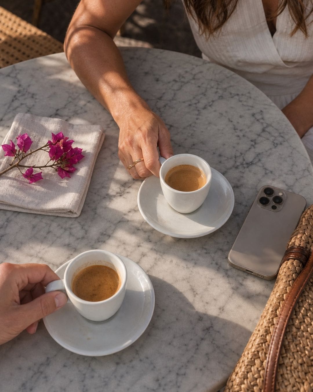 Two people share espresso cups at a marble table, with a pink flower napkin, a smartphone, and a woven bag nearby.